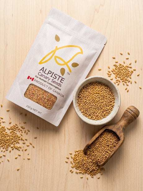 A stand-up pouch of Alpiste Canary Seeds for human consumption, styled on a light wood background with a small bowl, wooden scoop, and scattered seeds.