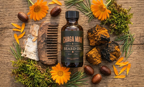 Chaga Man Beard Oil in an amber glass bottle with a black cap, styled on a rustic wooden background with orange flowers, acorns, moss, birch bark, and a wooden comb.